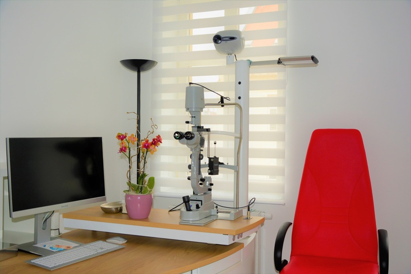 Ansicht der Augenarztpraxis: Ein Schreibtisch mit Bildschirm und Untersuchtungsgeräten, daneben ein Stuhl.